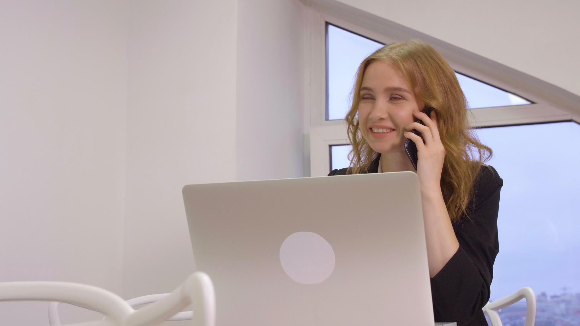 在办公室用笔记本电脑工作时微笑的商务妇女通过手机打电话视频的预览图