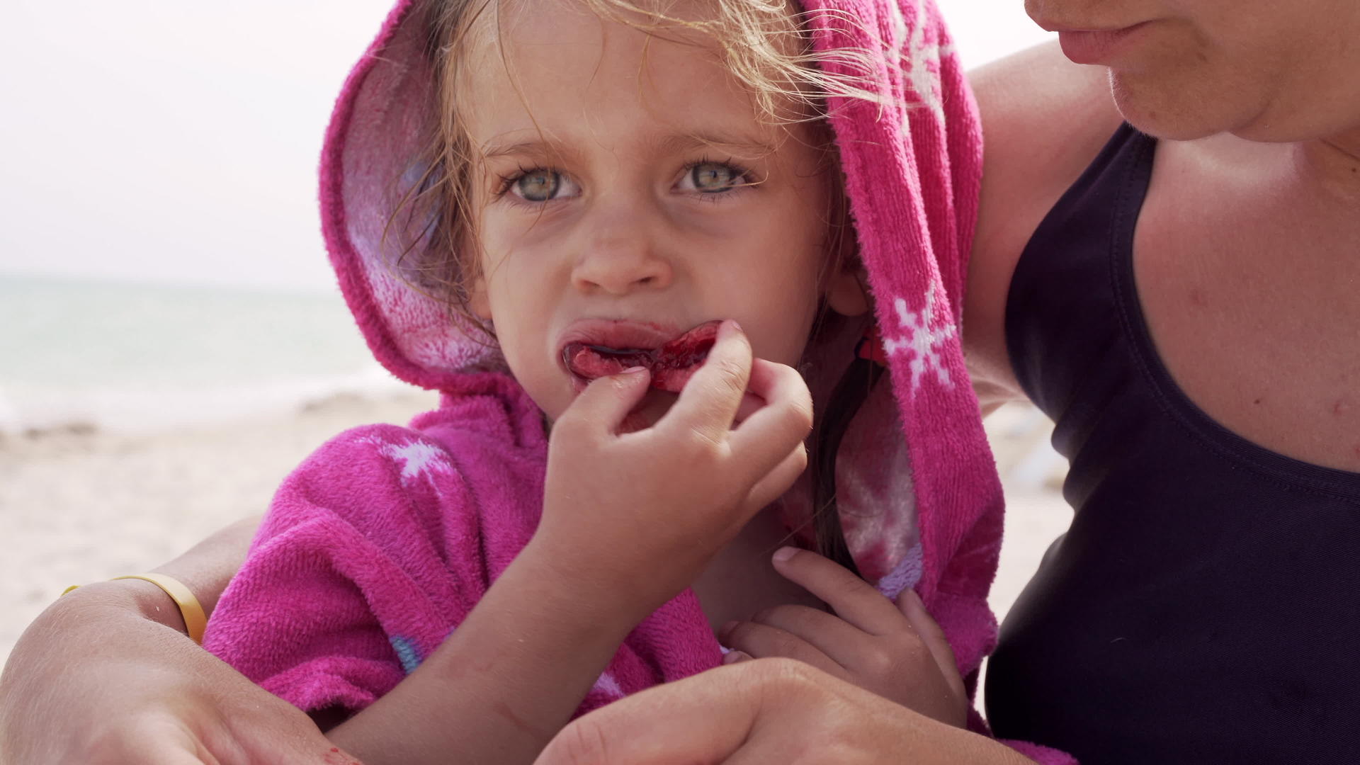 小女孩一边坐在妈妈的怀里一边吃着裹着沙滩毛巾的樱桃视频的预览图