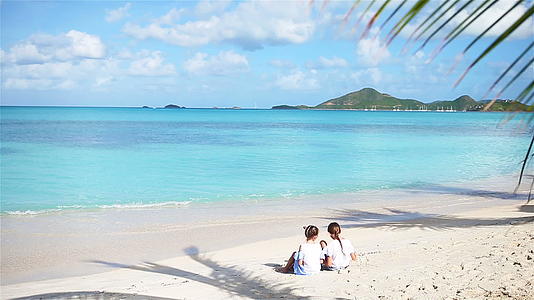 海滩上的两个小女孩快乐地坐在热带海滩的棕榈树上视频的预览图