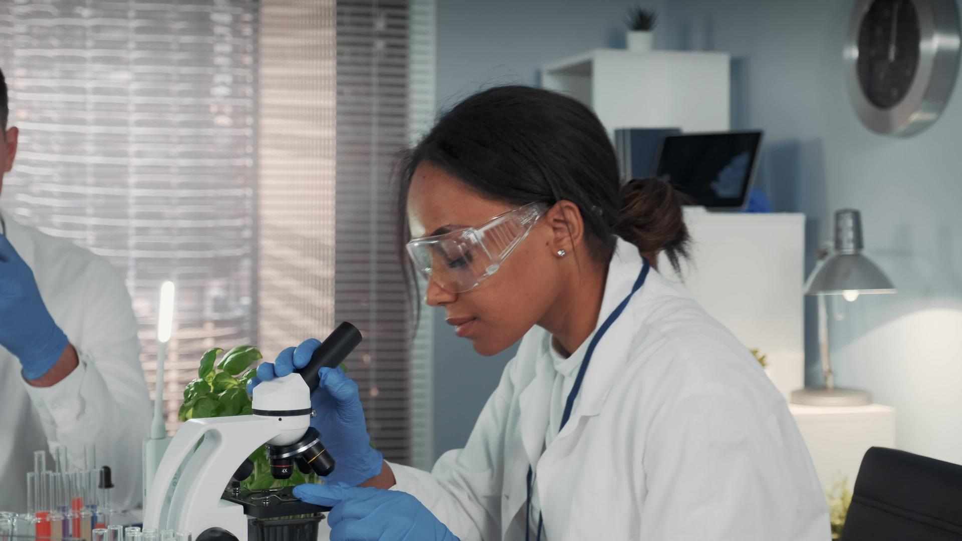 混血种族妇女穿着实验室外套和安全眼镜用显微镜工作视频的预览图