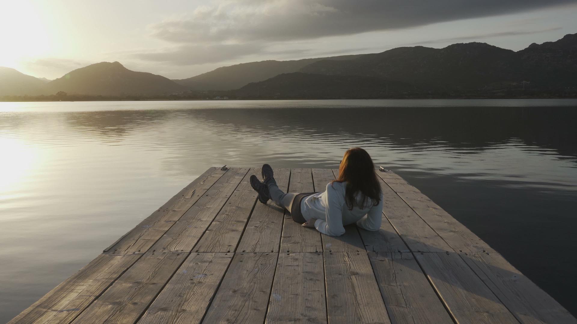 从后面看那个留着长发的年轻女孩日落时仰望着湖面躺在一个老旧的地方视频的预览图