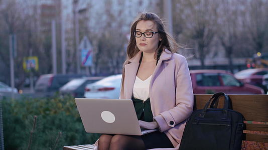 女性在笔记本电脑上打字坐在城市公园的长椅上视频的预览图