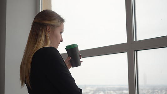 年轻手持咖啡杯的年轻女性设计师在窗口的背景下喝酒视频的预览图