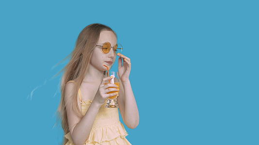 年轻的女孩用吸管从工作室的蓝色墙上的玻璃上喝橙汁女孩和青少年视频的预览图