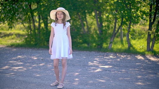穿着白色连衣裙和帽子的年轻女孩站在公园小巷的绿色树木背景下视频的预览图