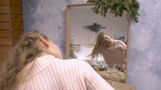 一个穿着粉色毛衣的年轻女孩在镜子前玩耍留着长卷发的圣诞树枝视频的预览图