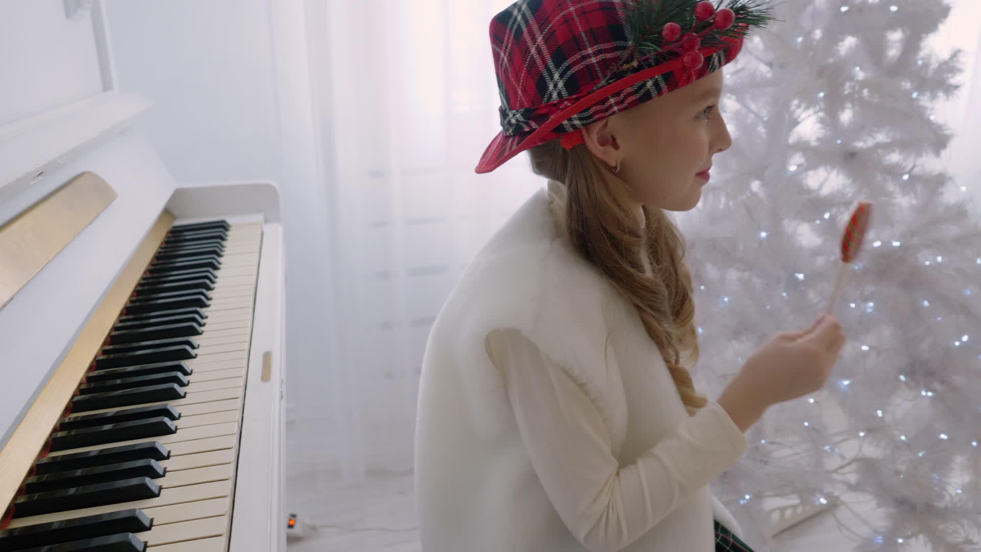 戴着红帽子的小女孩舔着白色钢琴和圣诞树旁的棒棒糖视频的预览图