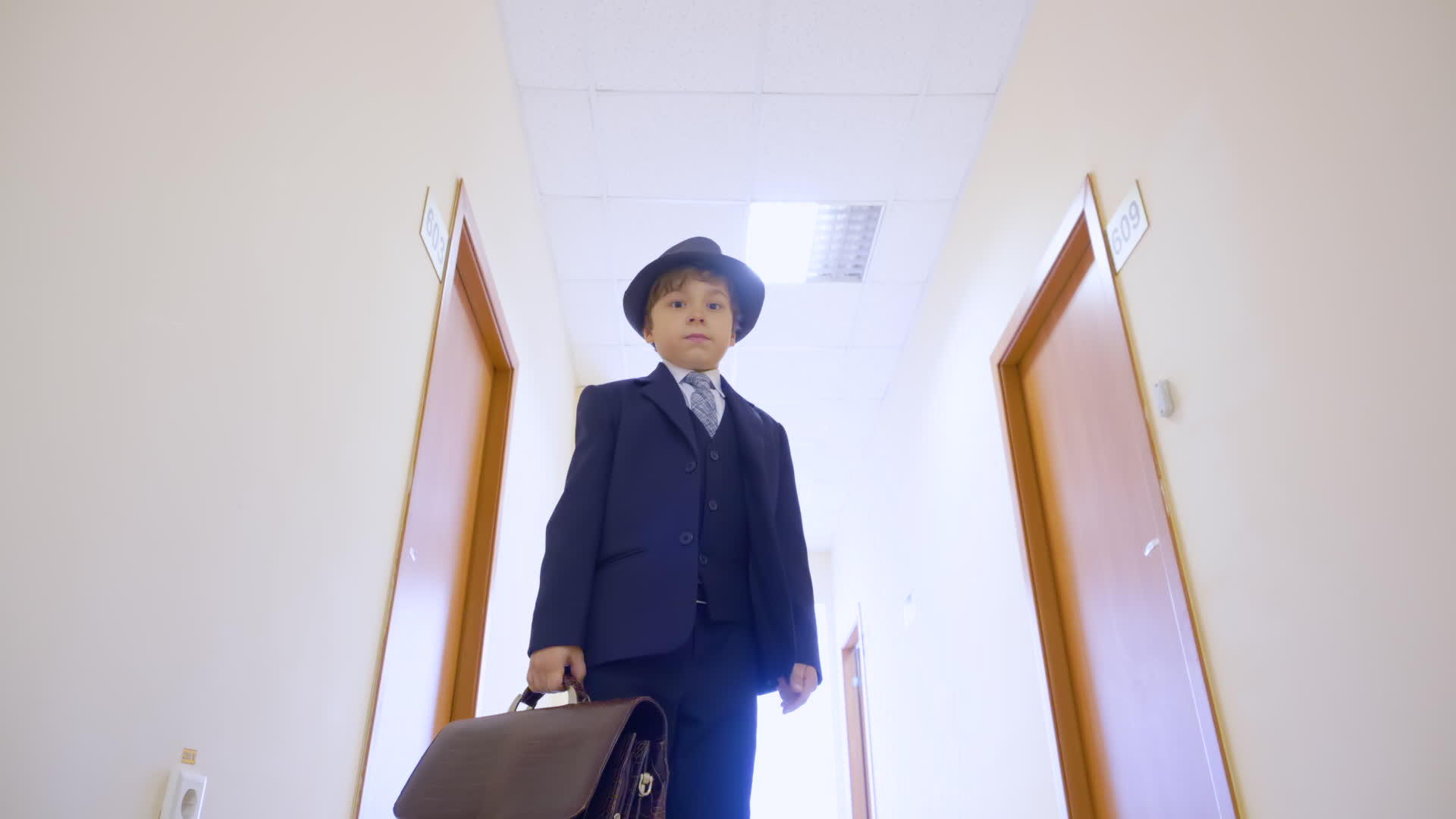 穿着西装的小男孩戴着帽子站在学校走廊里低角度看着镜头视频的预览图
