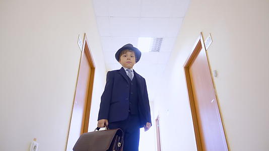 穿着西装的小男孩戴着帽子站在学校走廊里低角度看着镜头视频的预览图