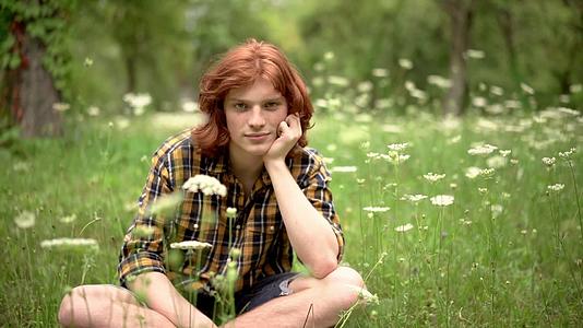 一个年轻的红发男孩坐在花园的草坪上长着野花视频的预览图