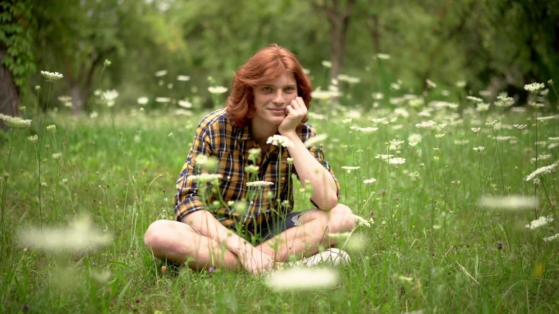 一个年轻的红发男孩坐在花园的草坪上长着野花视频的预览图