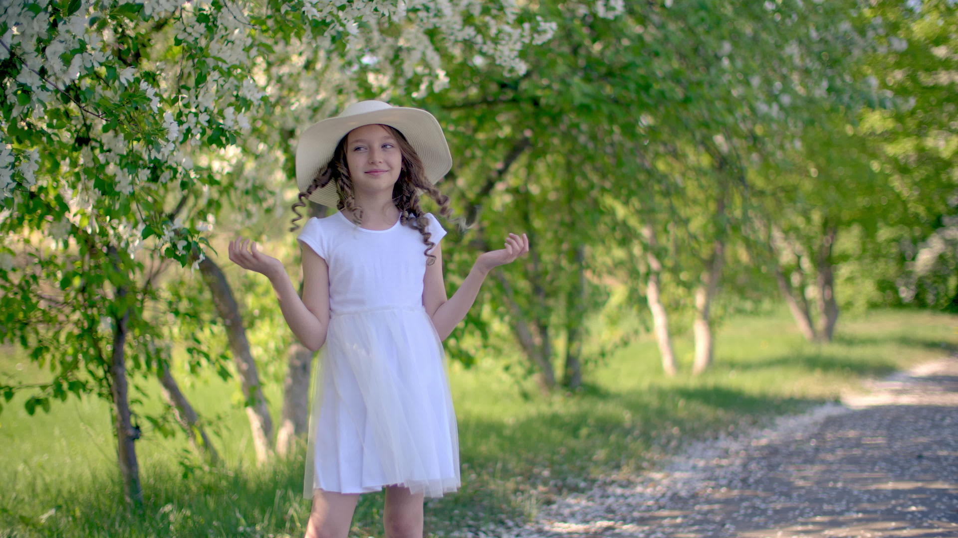 快乐的女孩戴着白色的帽子穿着春天花园里盛开的树木视频的预览图