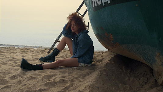 一个悲伤的女人坐在老船附近的沙滩上视频的预览图
