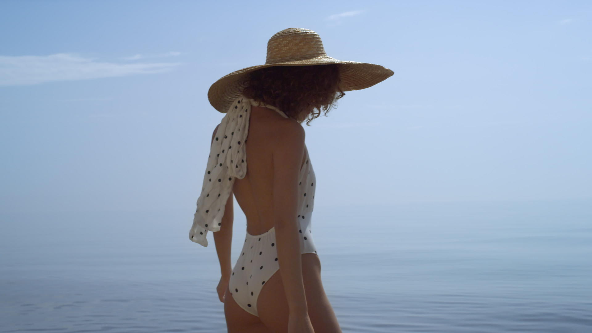豪华女士身穿宽边草帽走在蓝色海洋前的沙滩上视频的预览图