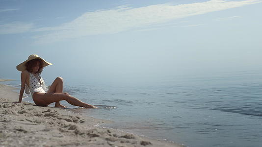 放松的女人坐在沙滩上用沙子在海浪中洗腿视频的预览图
