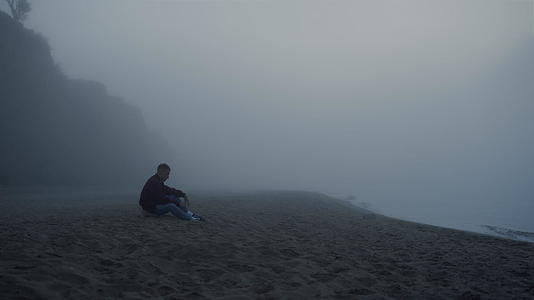 早晨在雾中坐在海边的沮丧的人视频的预览图