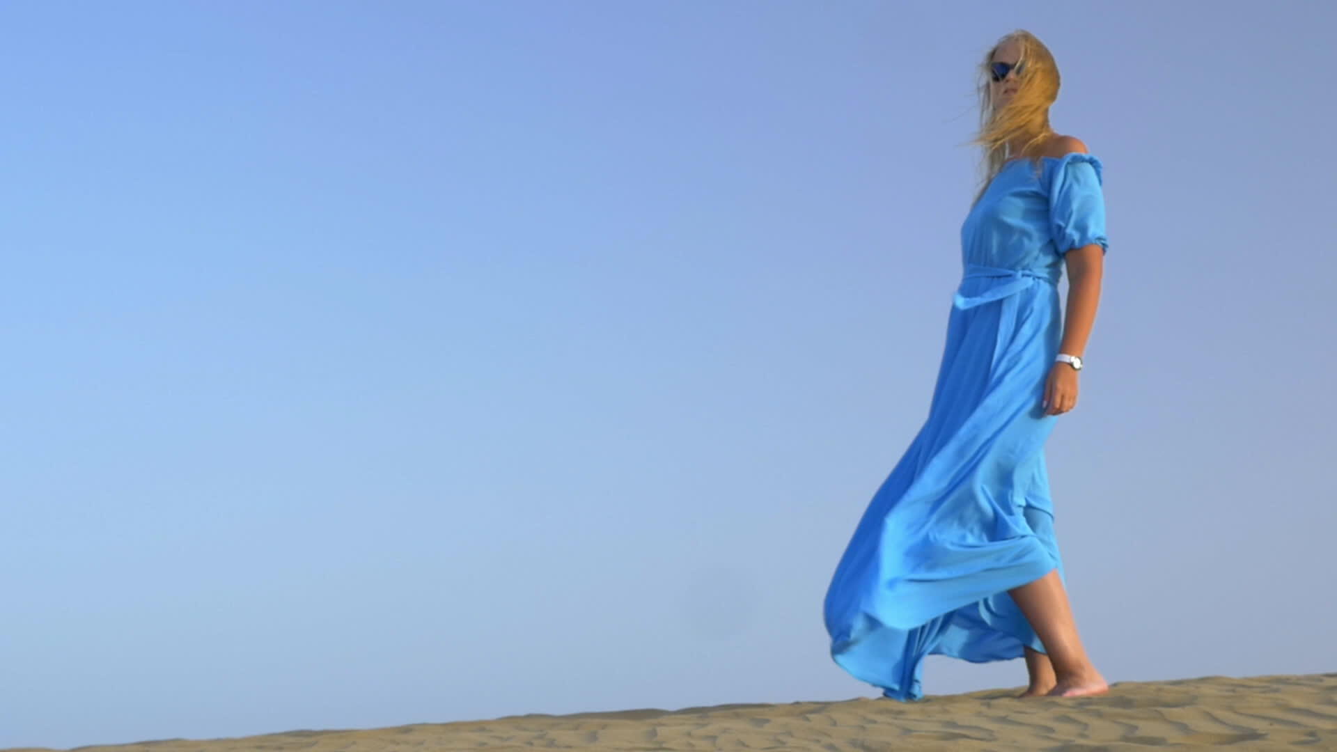 一个穿着蓝色长裙的年轻金发女子站在蓝天的慢动作视图中它在风中视频的预览图