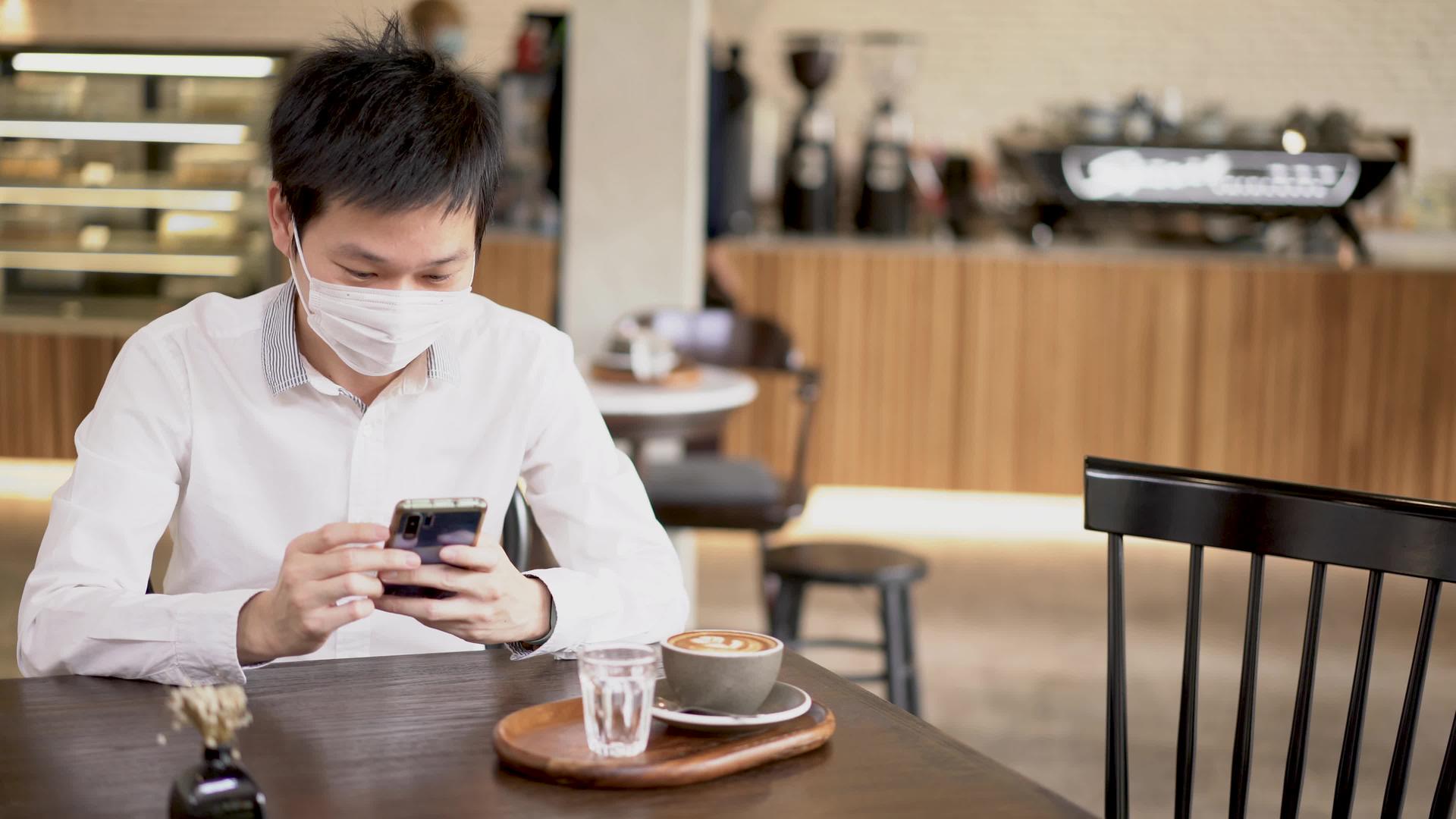 戴着外科口罩的亚洲男性使用智能手机在咖啡店订购饮料视频的预览图