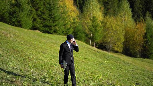 一个拿着笔记本电脑和黑色西装的男人走在山上在山上打电话视频的预览图