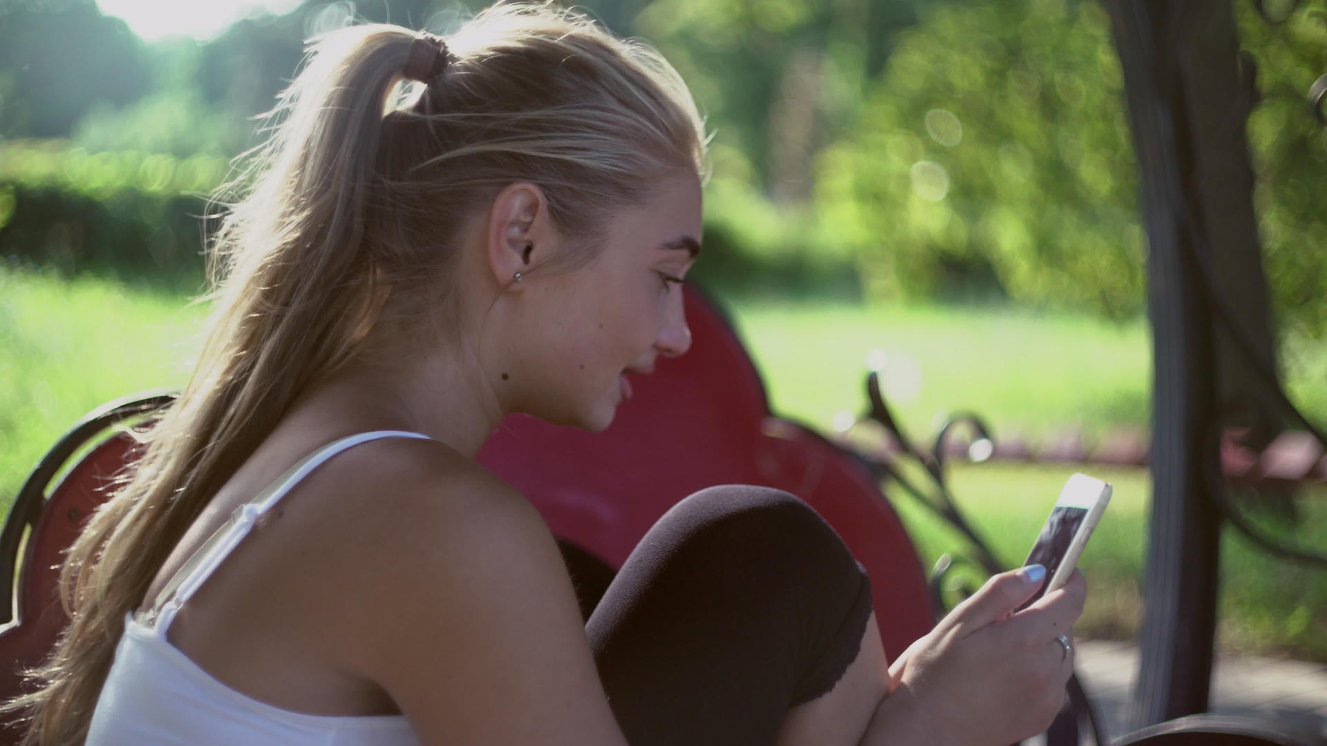 美丽的金发女孩坐在公园的长椅上微笑着查看手机视频的预览图