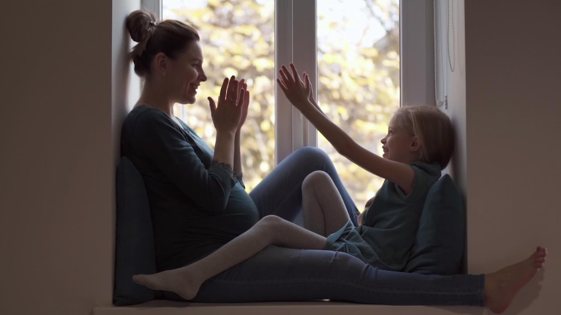 穿着绿松石的年轻孕妇和小女孩的女儿在窗台上鼓掌视频的预览图