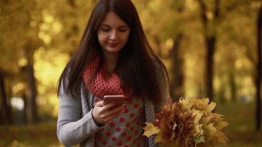 秋天女孩在公园里散步在她的智能手机上使用应用视频视频的预览图