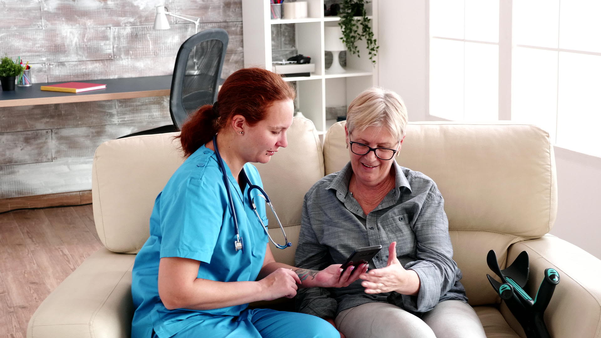 疗养院护理人员坐在沙发上帮助老年妇女使用智能手机视频的预览图