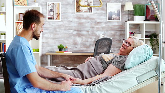 男性照顾者与躺在医院床上的生病老太太交谈视频的预览图
