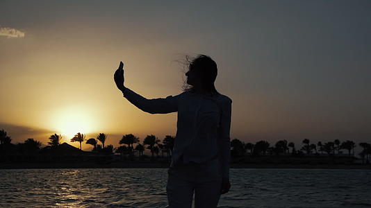 美丽的女人在日落的海边用手机自拍背光背影视频的预览图
