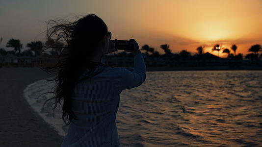 美丽的女人用手机拍摄日落海景视频的预览图