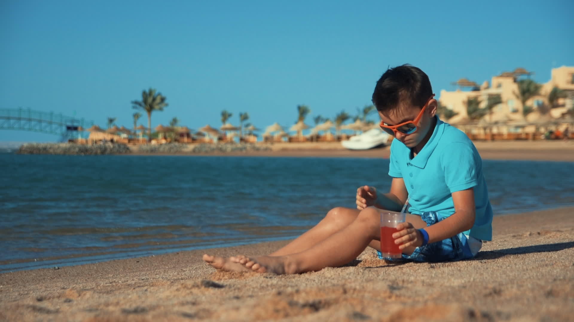 小男孩戴着墨镜在海滩上喝柠檬水玩手机视频的预览图