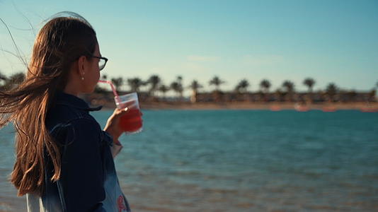 留着长发的小女孩在海边喝果汁享受美丽的风景视频的预览图