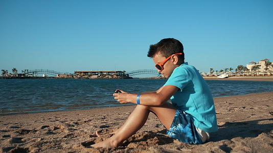 戴着太阳镜的小男孩在沙滩上玩手机视频的预览图