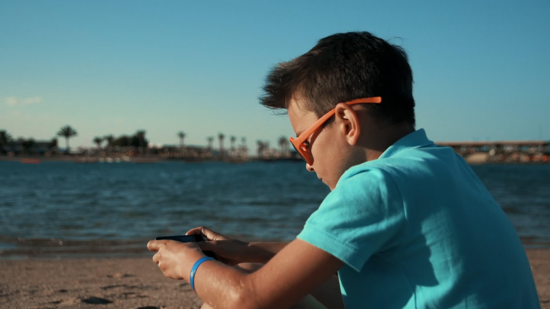 小男孩穿着蓝色T恤坐在海湾里用手机玩游戏视频的预览图