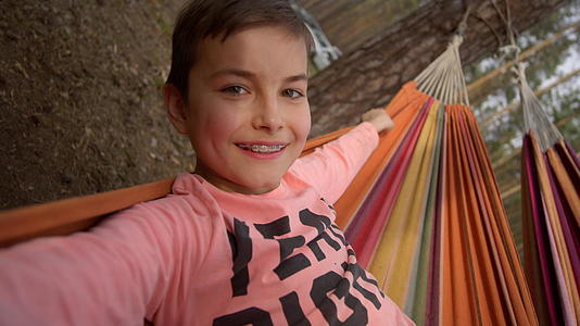 小男孩高兴地面对着镜头躺在秋天的树林吊床上享受时间和面部特写视频的预览图