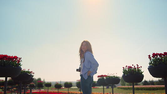 微笑的女孩穿着牛仔裤站在阳光下绽放的春天公园里拿着相机视频的预览图