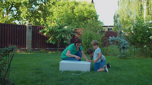 在阳光明媚的日子里年轻的母亲和两个孩子在后院打开了大纸箱视频的预览图