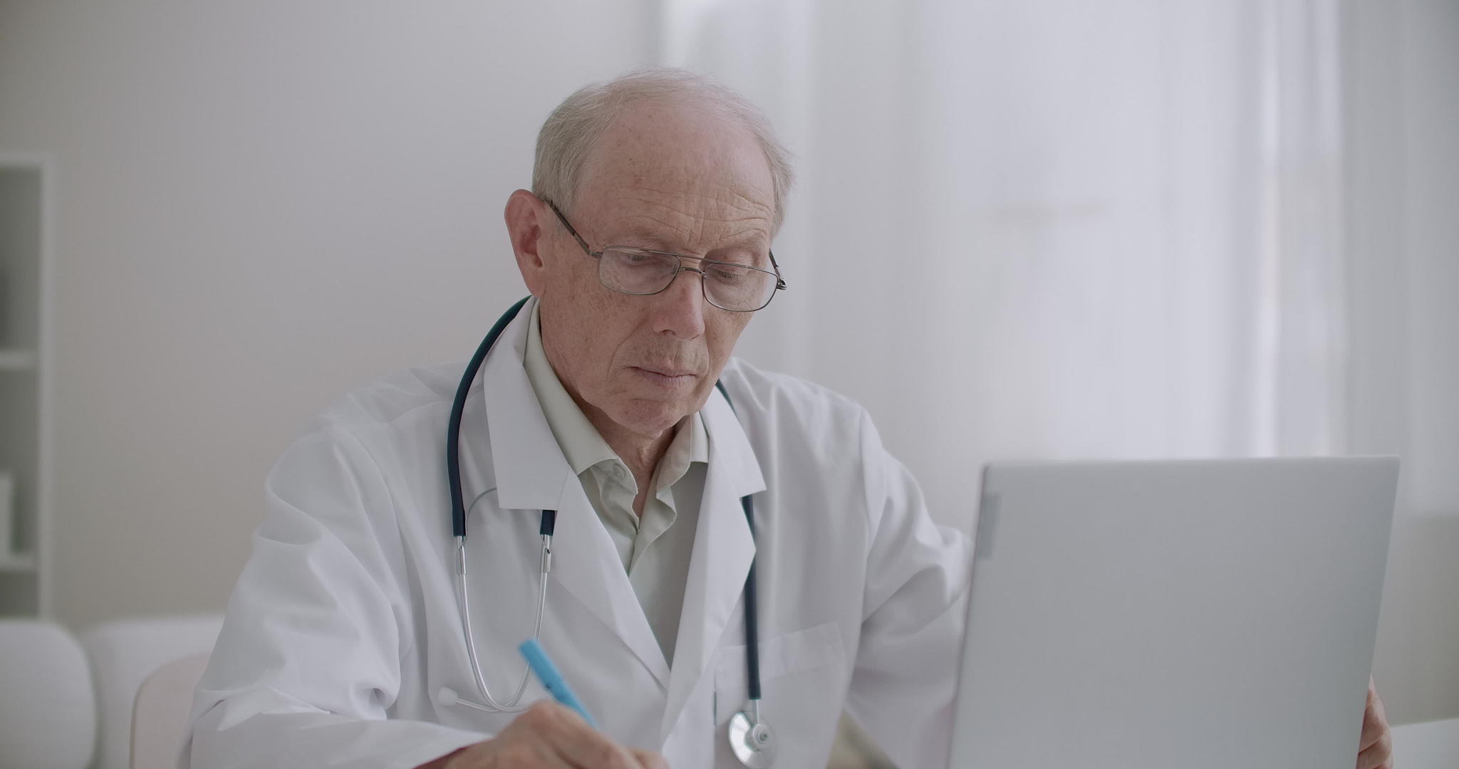 老年医学教授正在诊所办公室用膝电脑观看在线研讨会视频的预览图