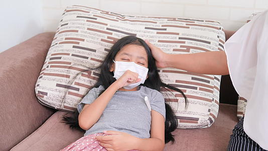 亚洲小女孩戴着外科口罩在沙发上咳嗽和生病妇女使用儿童视频的预览图