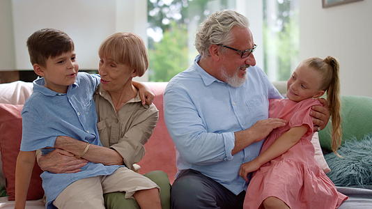 快乐的祖父和祖母坐在沙发上坐在大腿上的孙子孙女视频的预览图