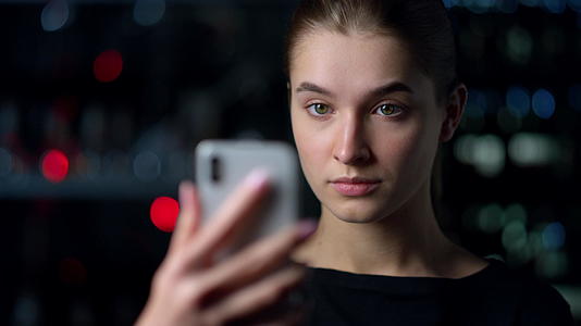 严肃的女孩用智能手机在夜间背景上自拍视频的预览图