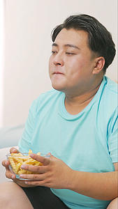 胖男生坐在沙发前边吃垃圾食品边看电视视频的预览图