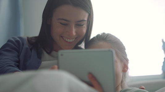 微笑的母亲陪着生病的女儿看平板电脑视频的预览图