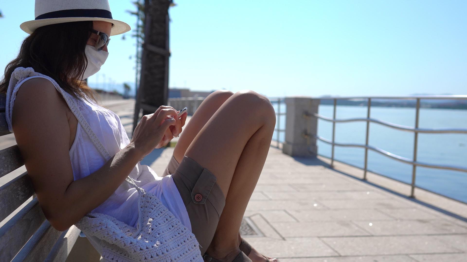 戴着白帽子的白人年轻女性坐在海边人行道上的智能手机长凳上视频的预览图