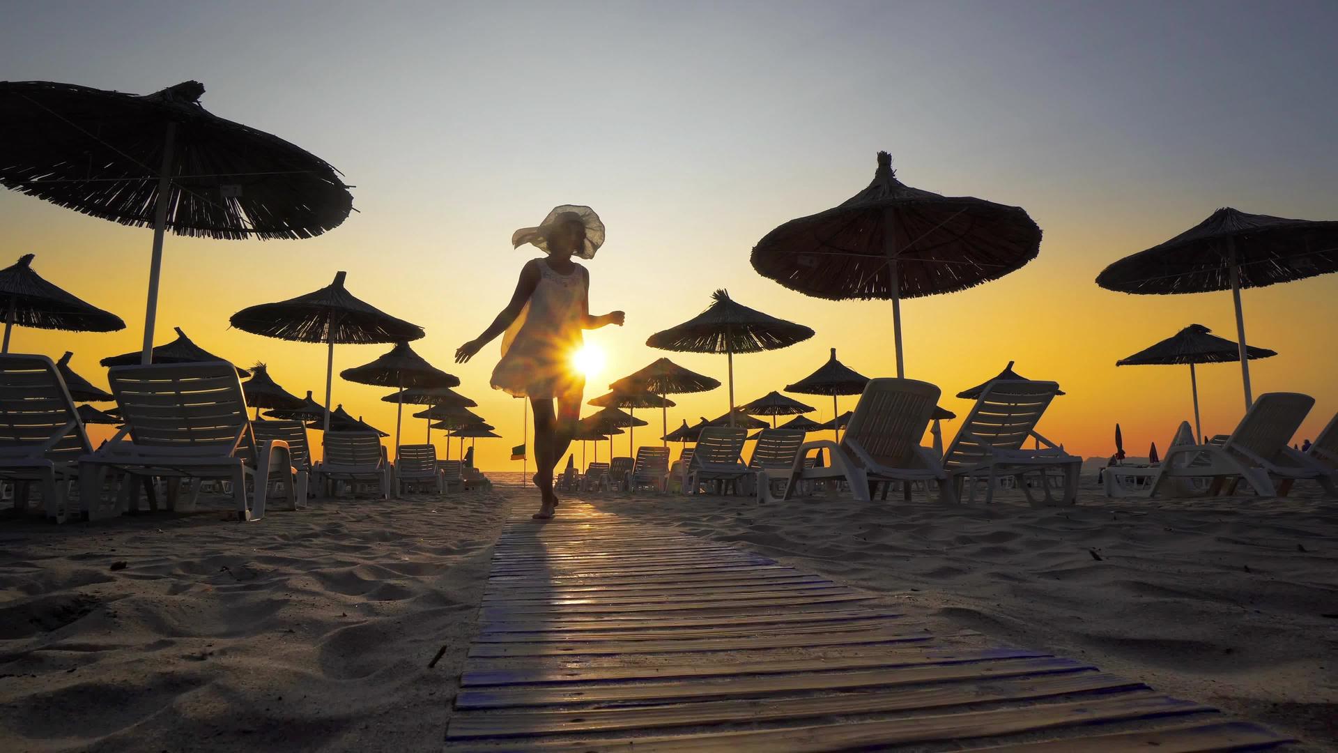 快乐的女人穿着白色的裙子戴着帽子日落时分在海滩上视频的预览图
