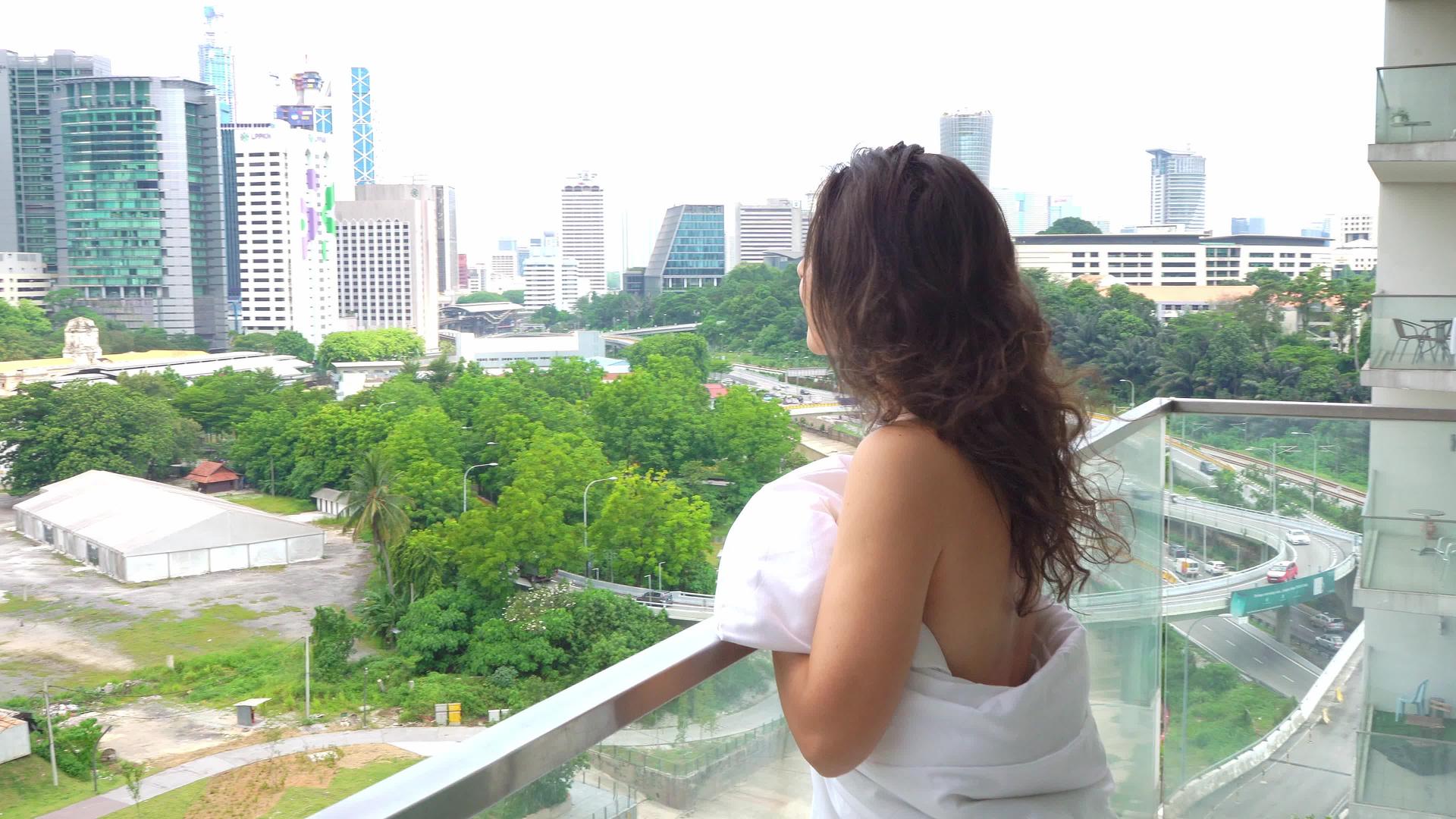 早上穿着白色毯子的女孩走到阳台上享受着现代大城市惊人的风景视频的预览图