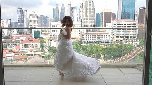 女孩穿着白色毯子在阳台上快乐地跳舞俯瞰现代城市视频的预览图