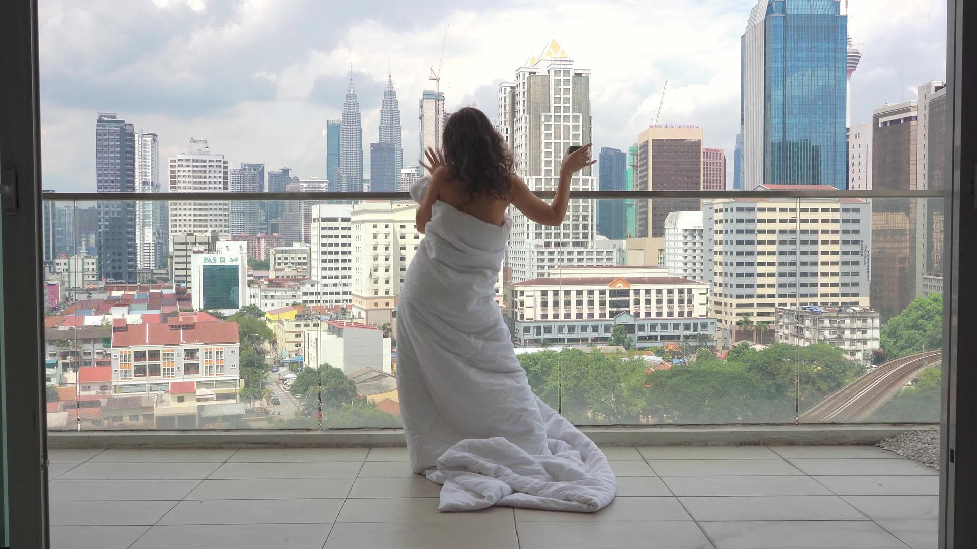 穿着白色毯子的女孩在阳台上快乐地跳舞俯瞰现代城市视频的预览图