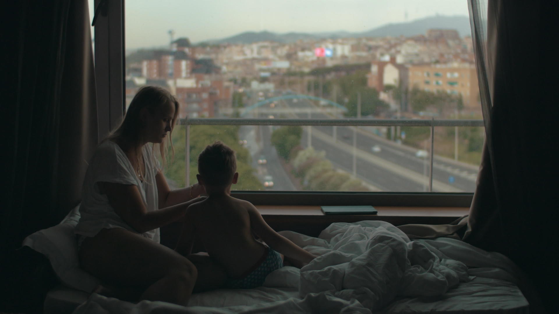 母亲和小儿子面对着巨大的全景窗城市风景躺在床上视频的预览图