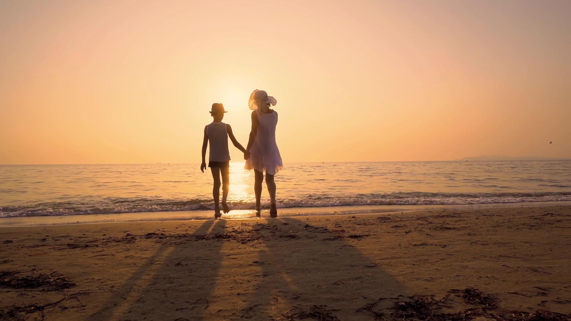 戴着帽子的母亲和儿子在空荡荡的海滩上走向日落的海岸冲浪视频的预览图
